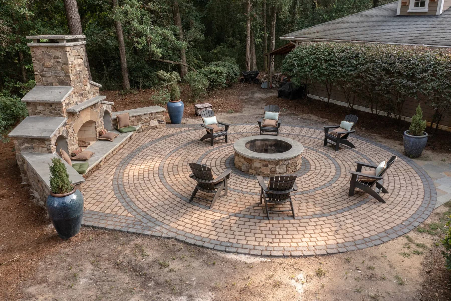 Aerial view of a circular paver patio with concentric borders, a central stone fire pit, Adirondack seating, and a large stone fireplace on James Island, SC